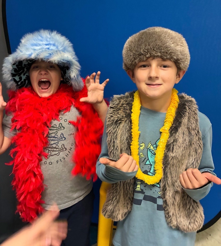 Two elementary students pose in front of a blue backdrop wearing furry hats, colorful leis, and a faux-fur vest. One makes a playful roaring expression.