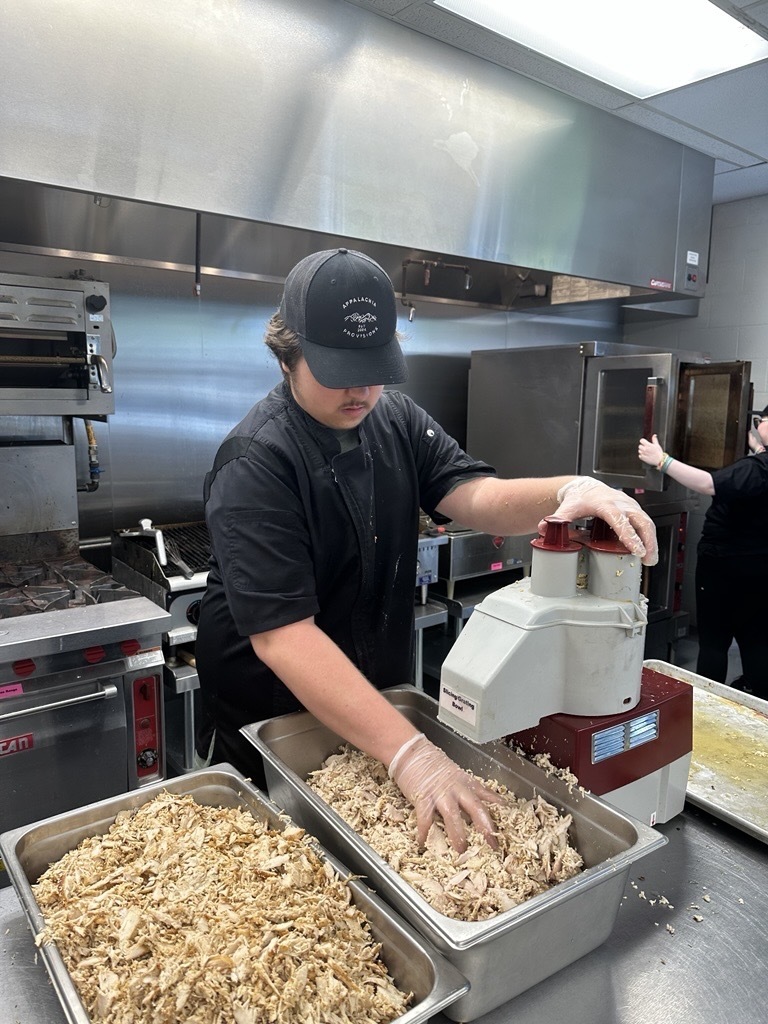 Student wearing an Appalachia Provisions chef uniform uses a commercial food processor to shred cooked chicken in the café kitchen.