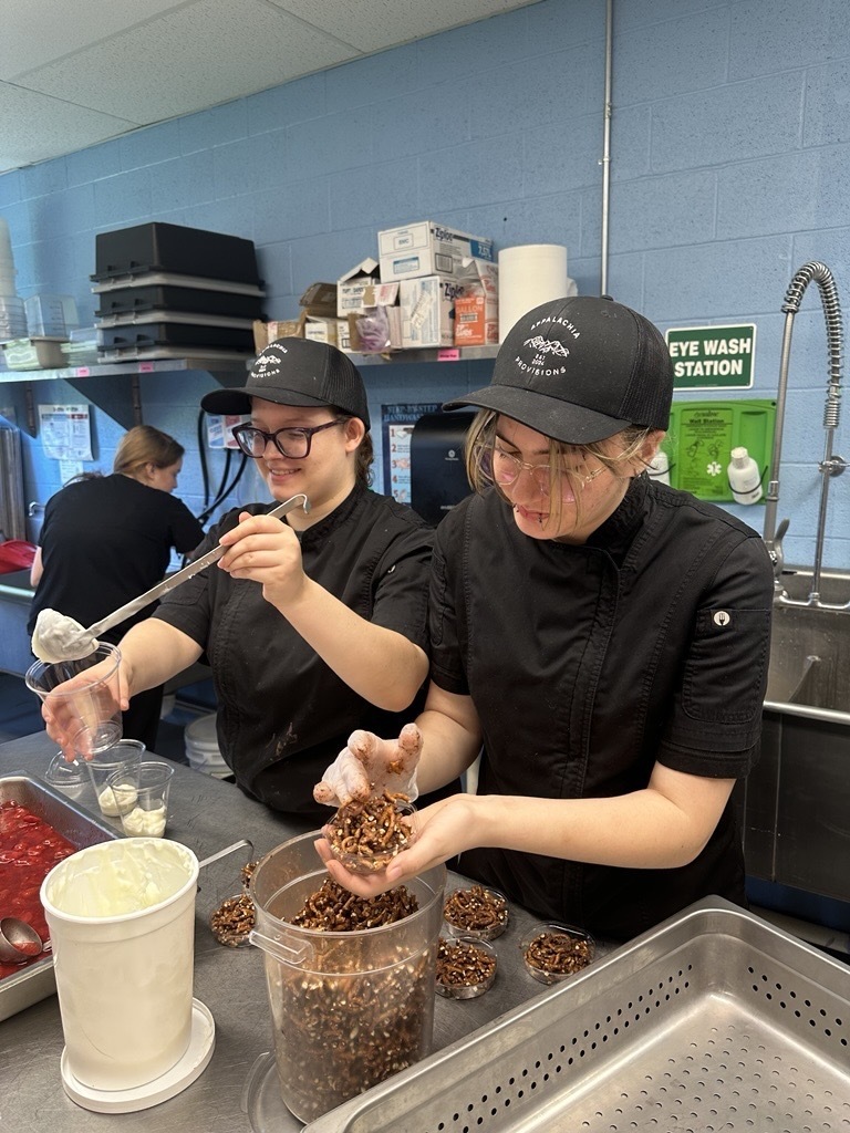 Two culinary students prepare yogurt parfaits, layering yogurt, strawberries, and chocolate-covered pretzels in clear cups.