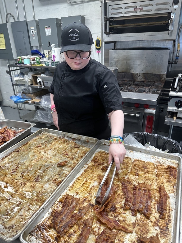 Student wearing an Appalachia Provisions chef uniform uses tongs to transfer cooked bacon from a baking sheet.