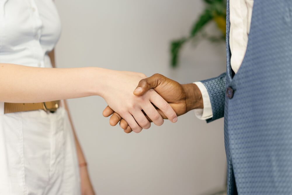 Image of a woman in a white shirt and pants shaking the hand of a man in a blue suit. The image focuses on the hands