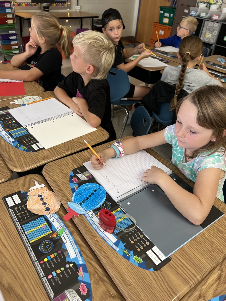Several students sit at desks writing in notebooks. A girl in the foreground is focused on her paper, with colored cut-out shapes on her desk.