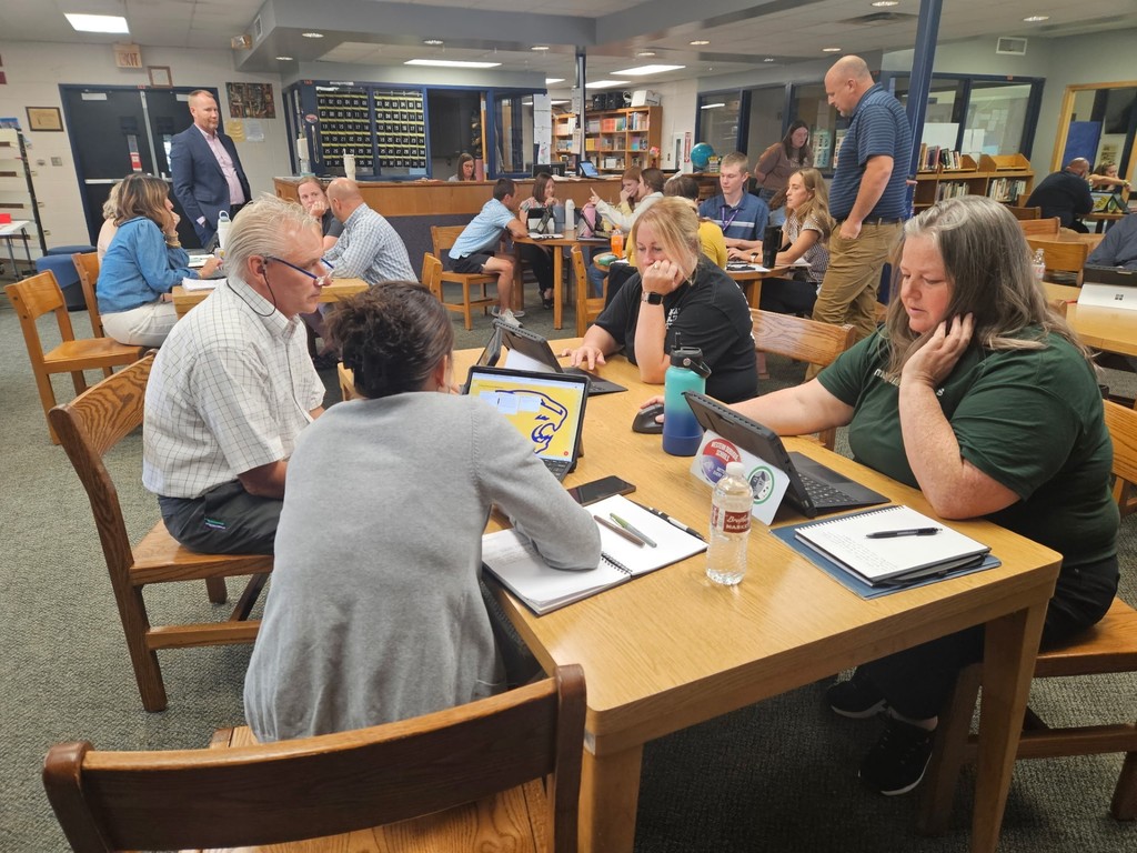 A group of adults seated around rectangular tables in a library, working with notebooks and laptops. Some participants are writing while others are looking at screens