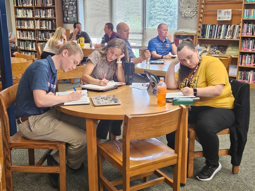 Three adults seated at a round wooden table in a library, writing in notebooks. They are focused on their work, with bookshelves and other small groups visible in the background.