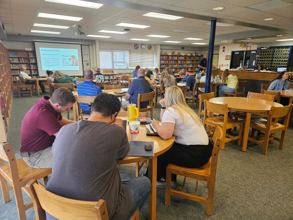 A library setting where multiple groups of adults sit at round tables. A projector screen at the front shows a slide titled “Whole Group Discussion,” while participants take notes and discuss in small groups.