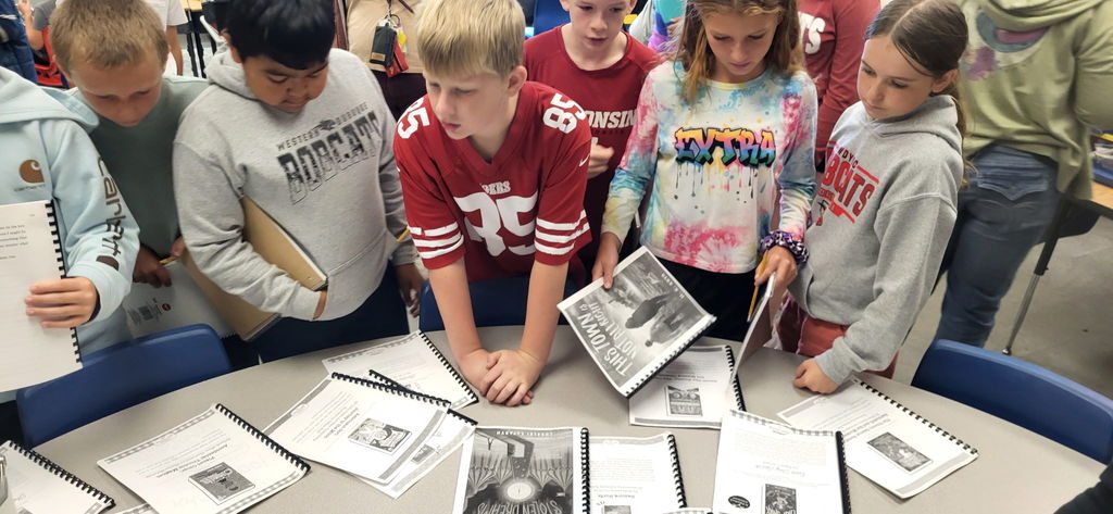 Group of students gathered around a table looking at spiral-bound booklets. Each holds or examines a reading packet together.