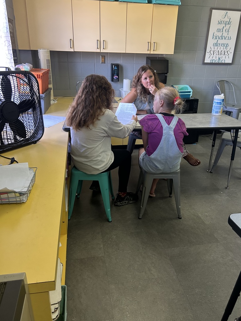 Teacher sits with two students at a small table, listening while a student reads aloud from a paper.