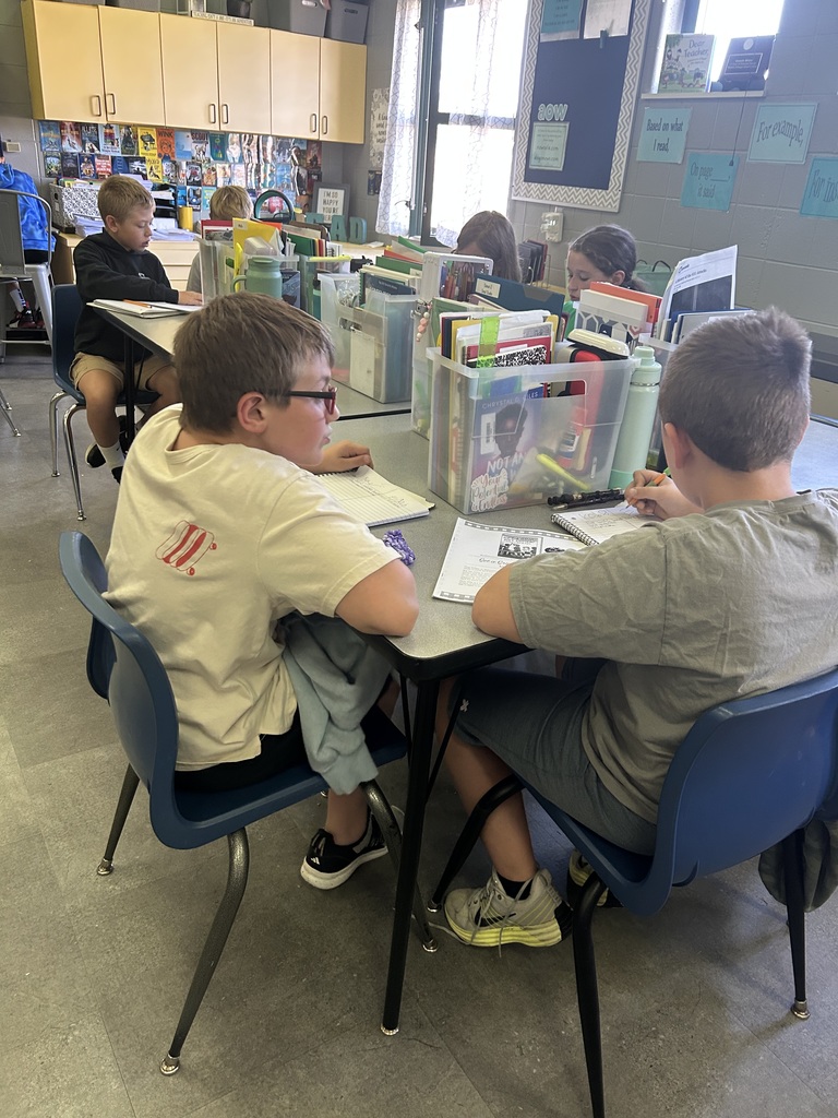 Students sit at tables with bins of books and supplies, writing in notebooks and working independently.
