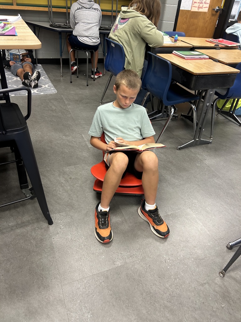 Student sits on a red stool reading a book while others nearby read at desks and on the carpet.