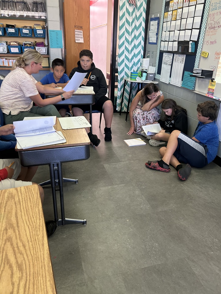 eacher works with two students at desks while a small group of students read and write together on the floor nearby.