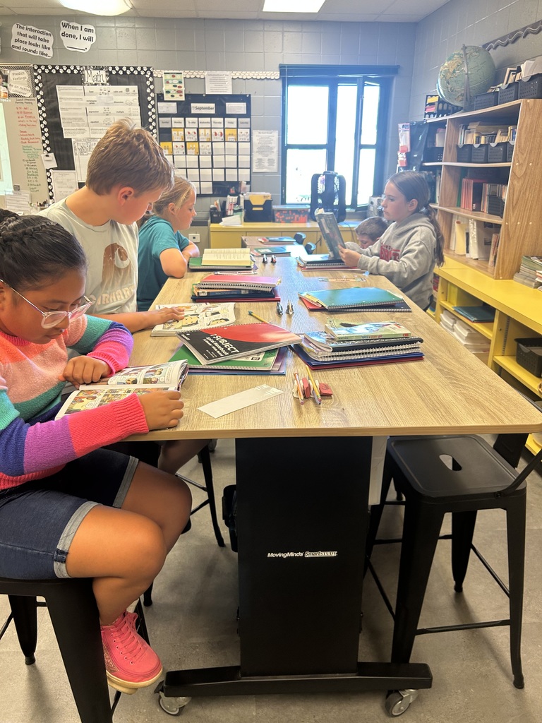Students at a rectangular table reading books quietly. Classroom shelves and supplies are visible in the background.