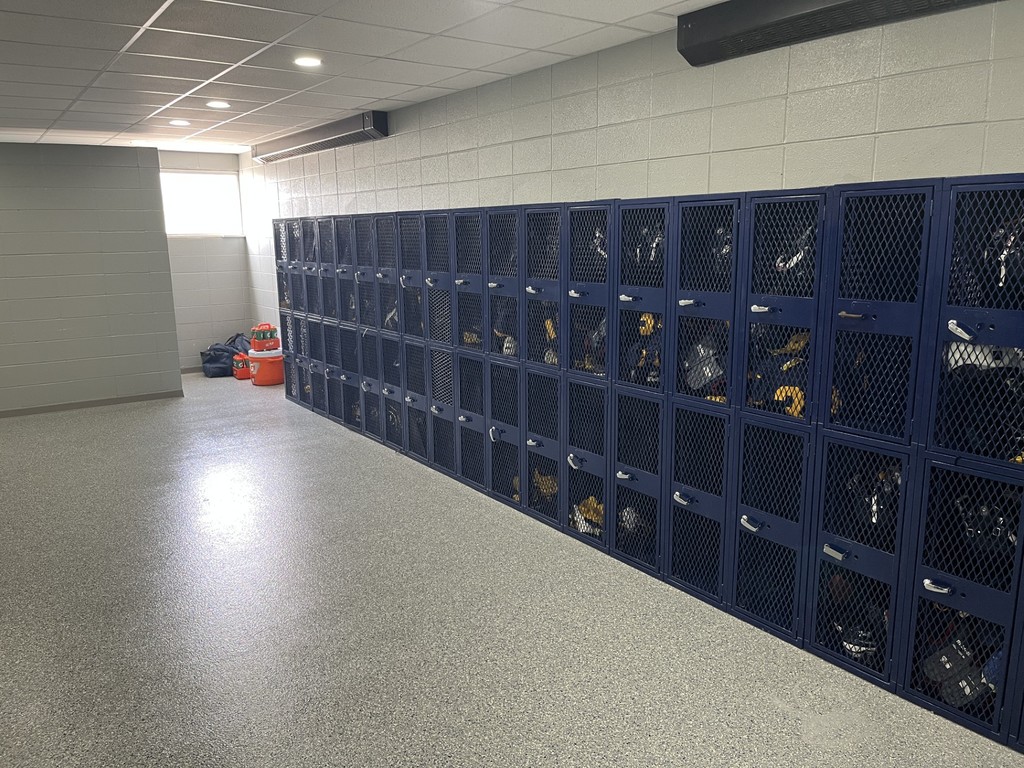 Locker room with a long row of blue mesh-front lockers containing athletic gear. Sports bags and orange water jugs are stacked near the window.