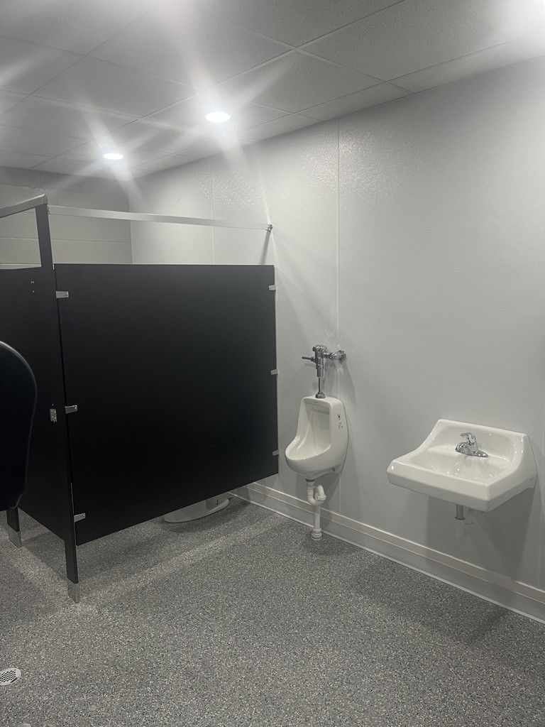 Restroom with a single stall divider and wall-mounted sink beneath bright ceiling lights.