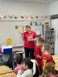 Teacher in a red shirt stands at the front of the classroom holding a bag of materials, speaking to students seated at desks and working on a project.