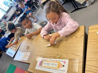 Young student in a pink sweatshirt works intently at a desk, folding or arranging craft sticks, while a classmate looks on.