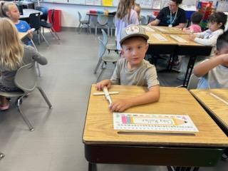 Student wearing a gray shirt and baseball cap sits at a desk holding craft sticks, with other children working nearby.