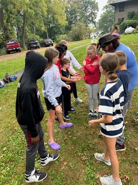 Students stand in a circle outside on the grass, listening closely to an adult leader giving instructions for an activity.