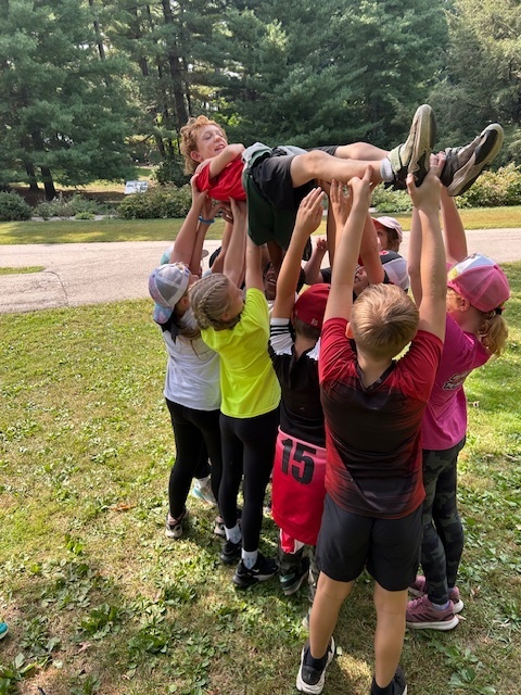 Students outdoors lift a teammate high in the air with their arms during a team-building trust exercise.