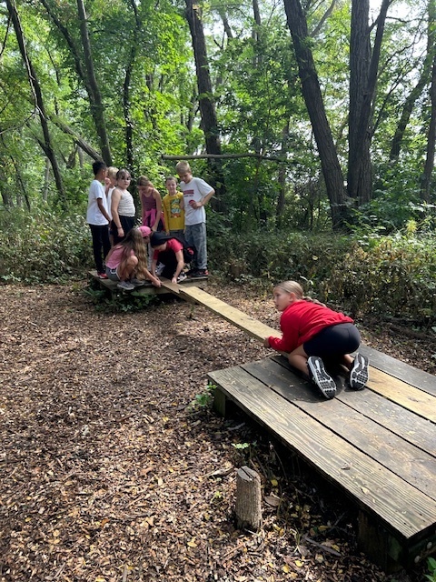 Group of students work together in the woods, using a wooden plank to cross from one platform to another.