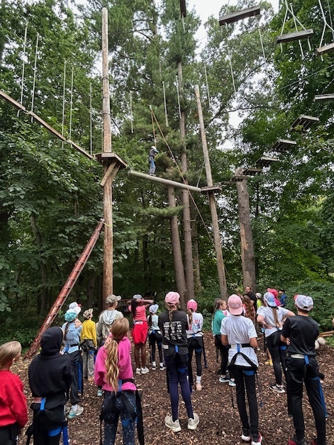 Large group of students wearing helmets and harnesses gather at the base of a ropes course, watching as one participant climbs high among the trees.