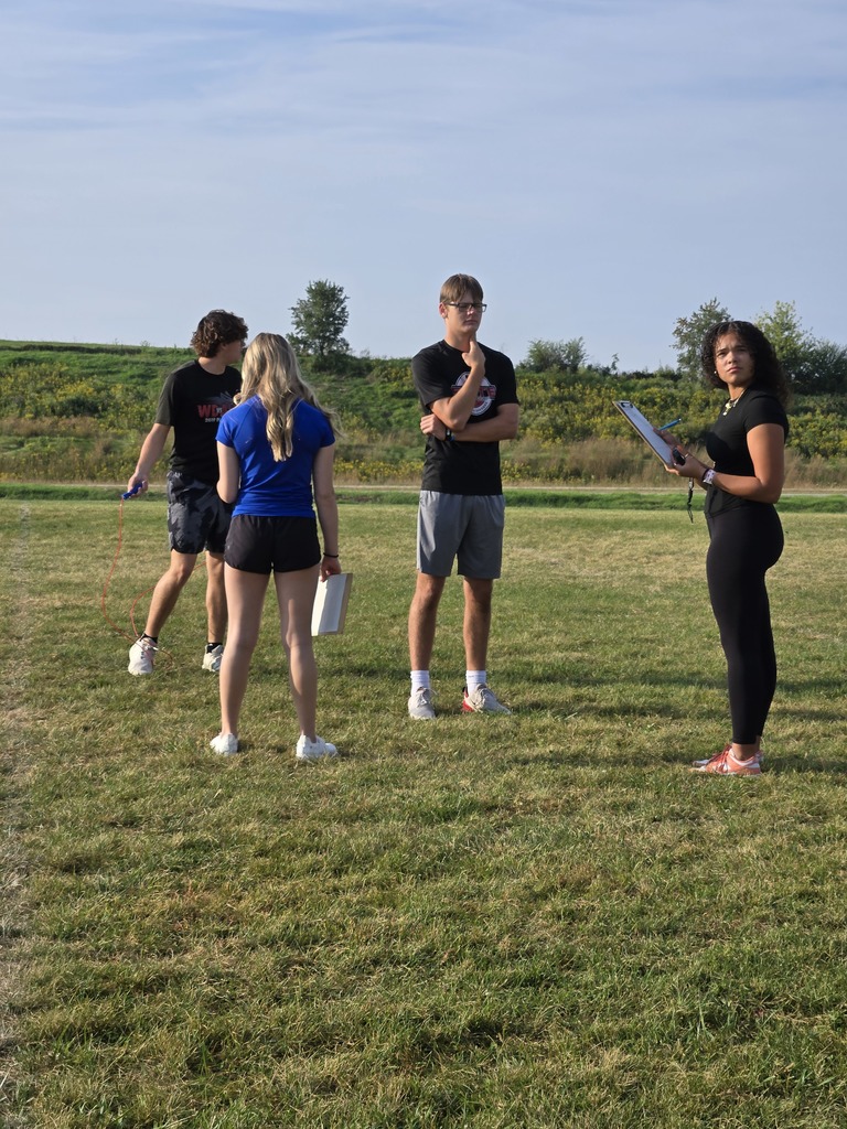 Small group of students stands together outdoors; one student holds a clipboard while others watch and discuss.