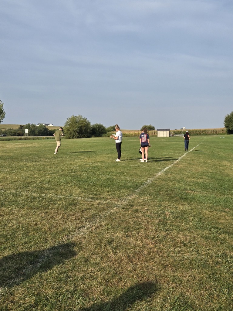 Students and a teacher stand spread out on a grassy field under a clear sky, preparing for an outdoor activity.