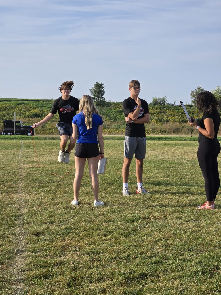 Student jumps rope while three others, including two holding clipboards, observe and record on a grassy field.