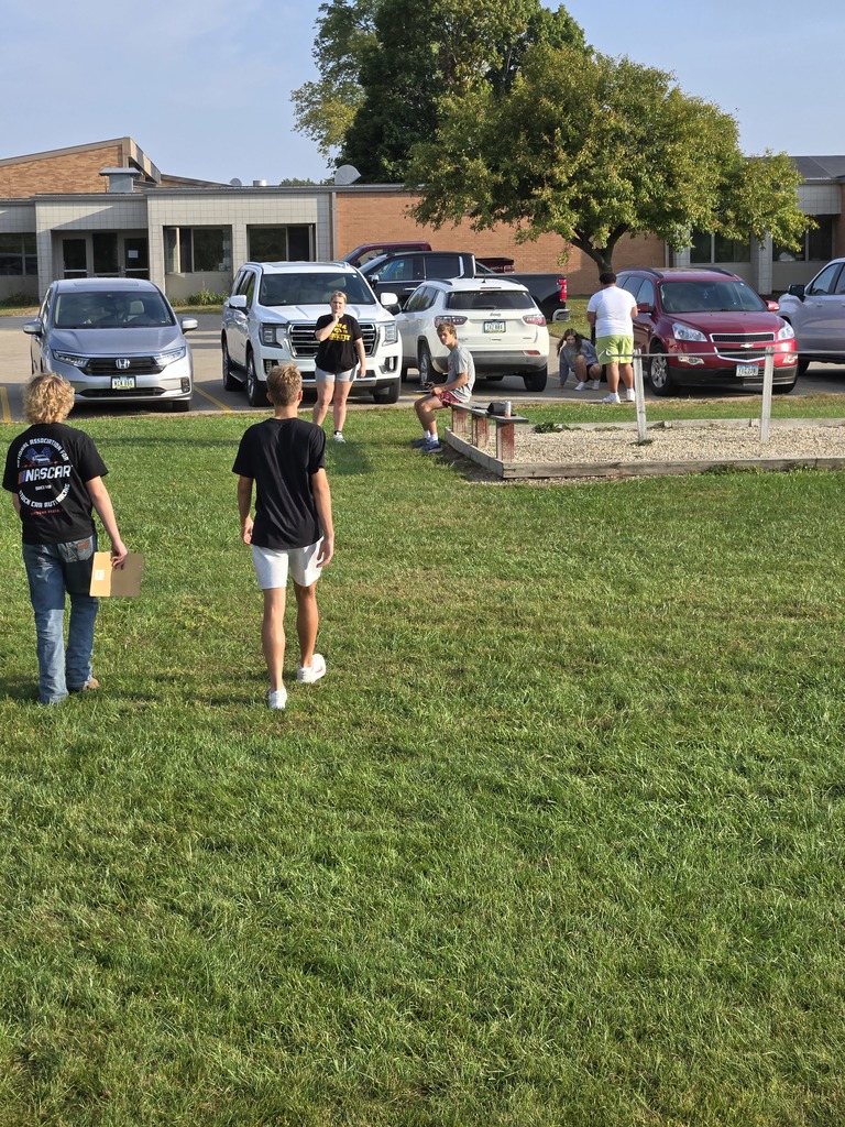 Students gather outside near parked cars and a school building, some standing and others sitting, during an outdoor class activity.