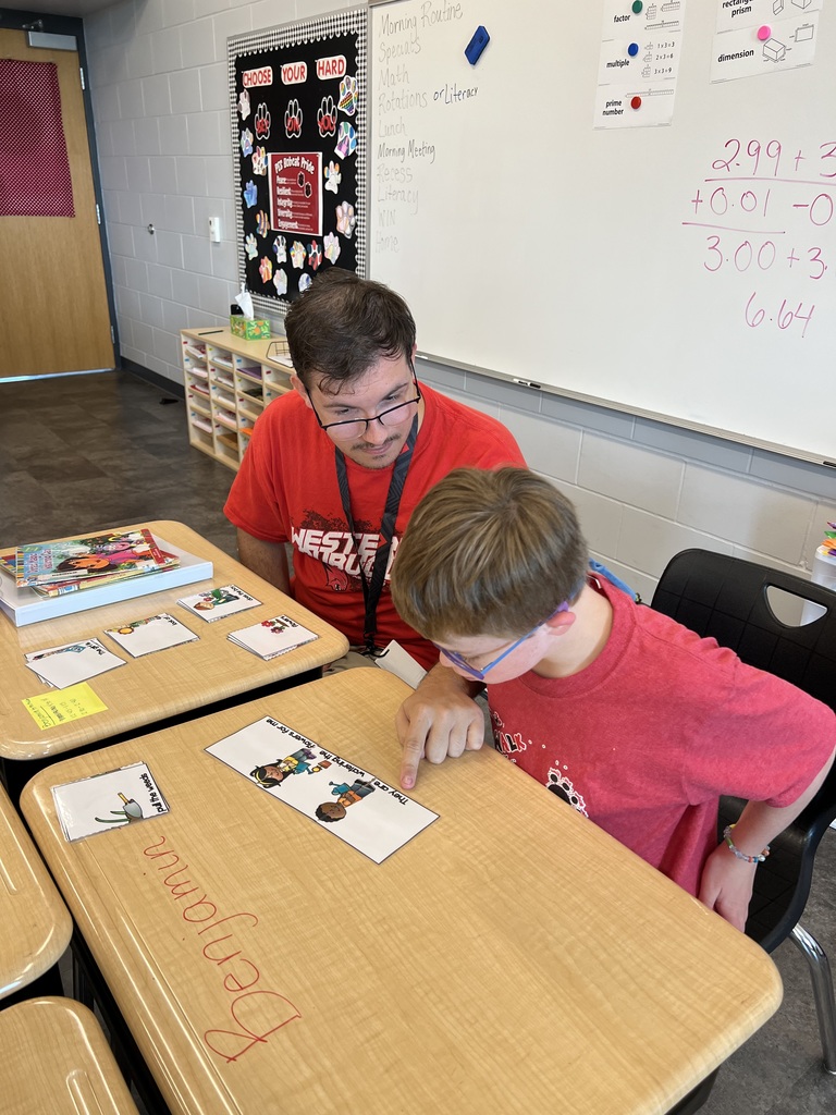 Teacher in a red shirt sits beside a student at a desk, guiding the student as they point to a reading activity card.