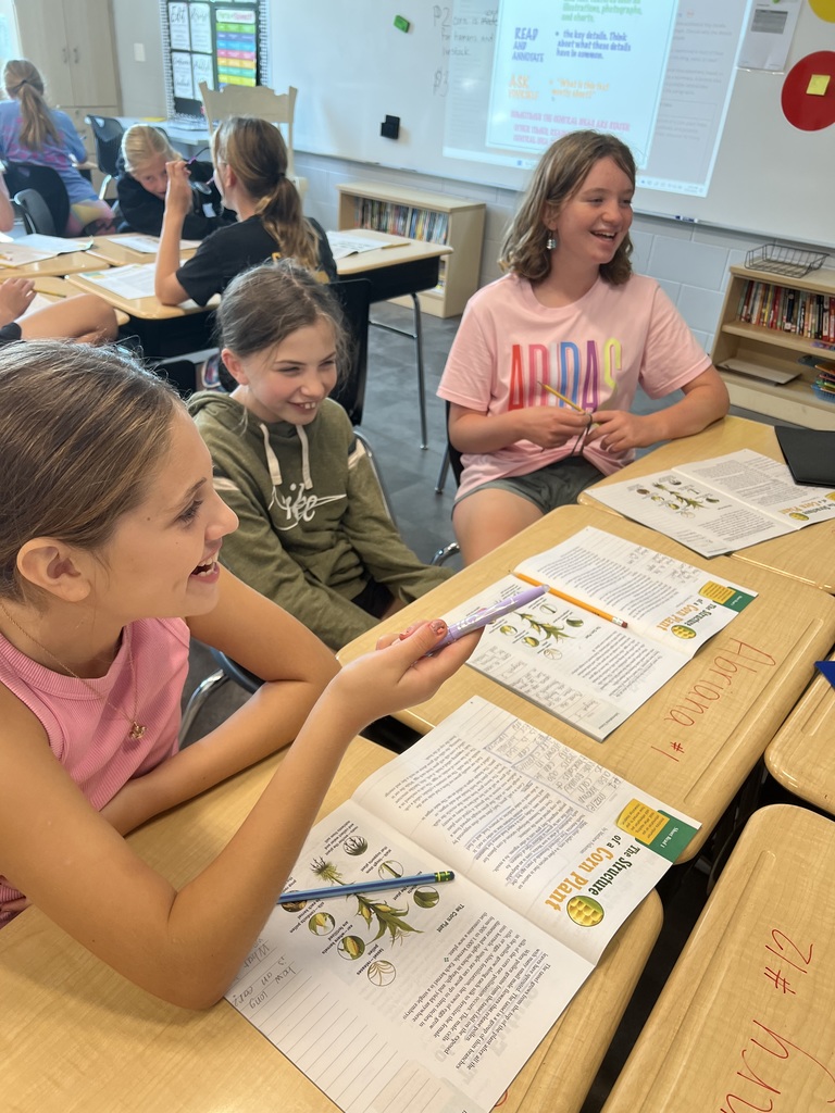 Three students sit at desks smiling and laughing during a classroom activity.