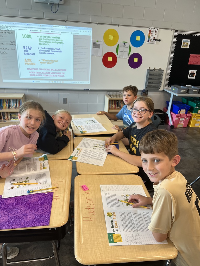 Six students sit together at two desks in a classroom, looking at the camera while working on assignments in open workbooks