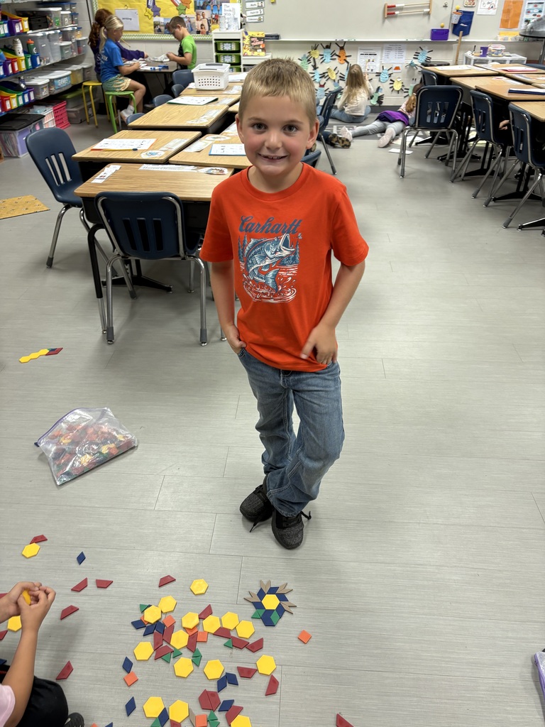 Student wearing an orange shirt stands in the classroom, smiling next to a pattern made from colorful geometric shapes spread on the floor.