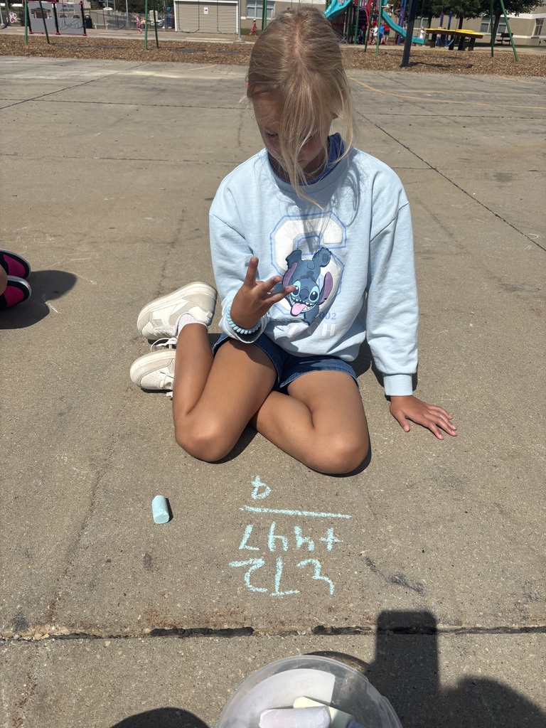 Student sits on the blacktop, counting on her fingers while solving a math problem written in blue chalk.