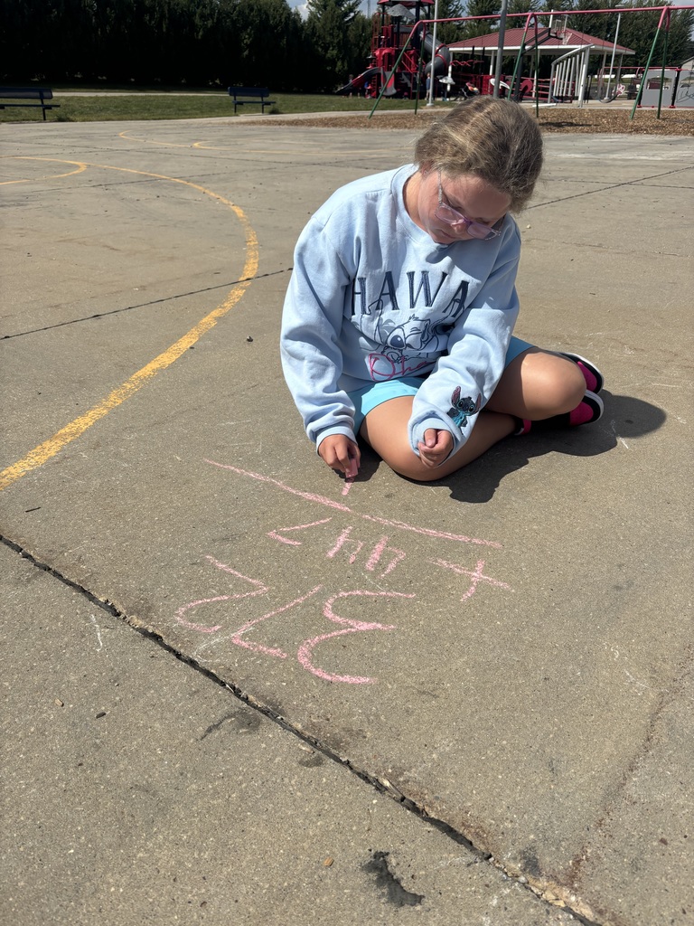 Student sits on the blacktop playground, writing a long division problem in pink chalk while concentrating on her work.