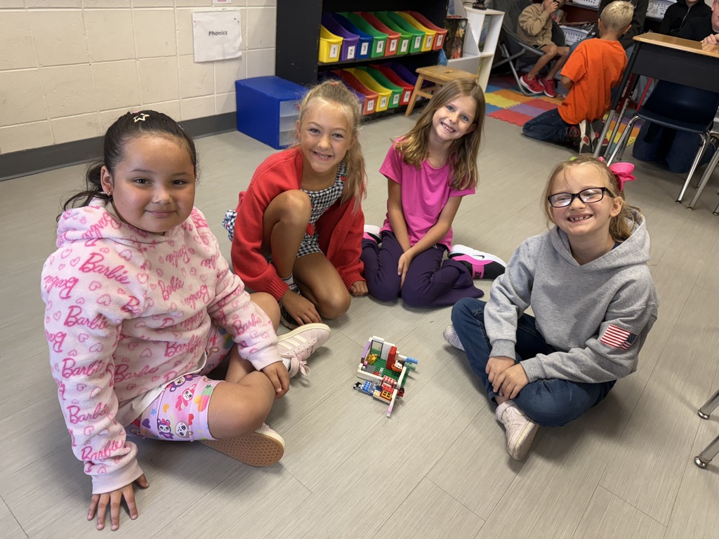 Four students sit on the classroom floor, smiling next to a small LEGO creation they built together.