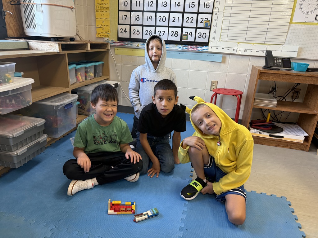 Four students on a classroom rug smile beside a LEGO structure they built together.