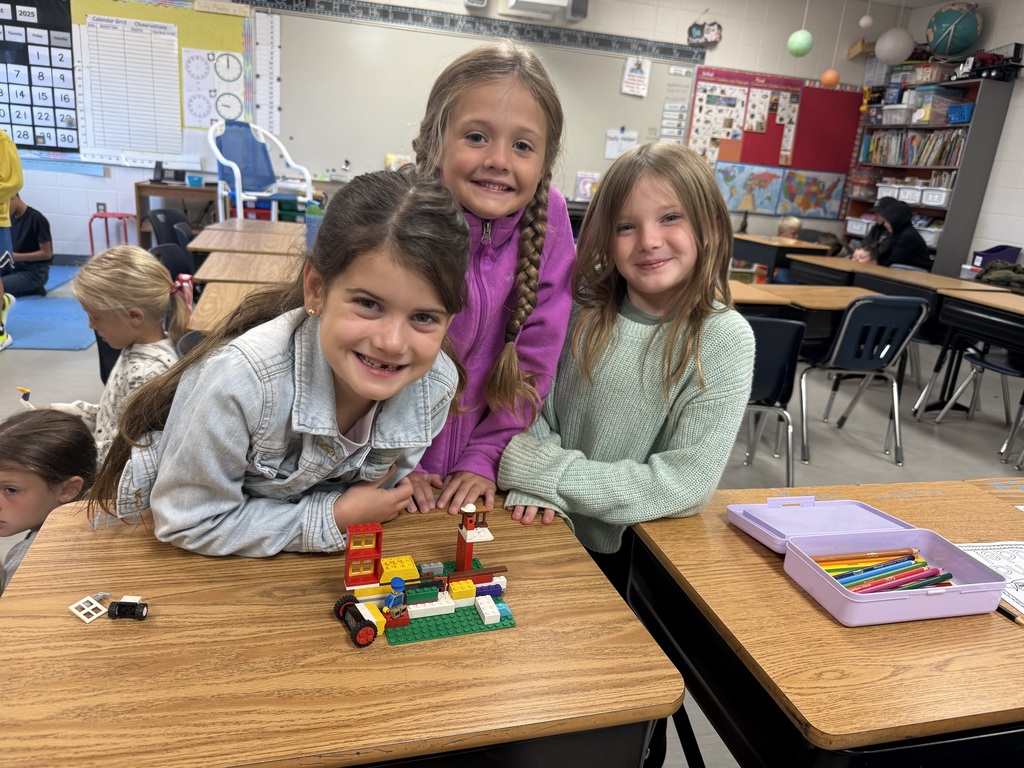 Three students at a desk lean in proudly to show their colorful LEGO build.