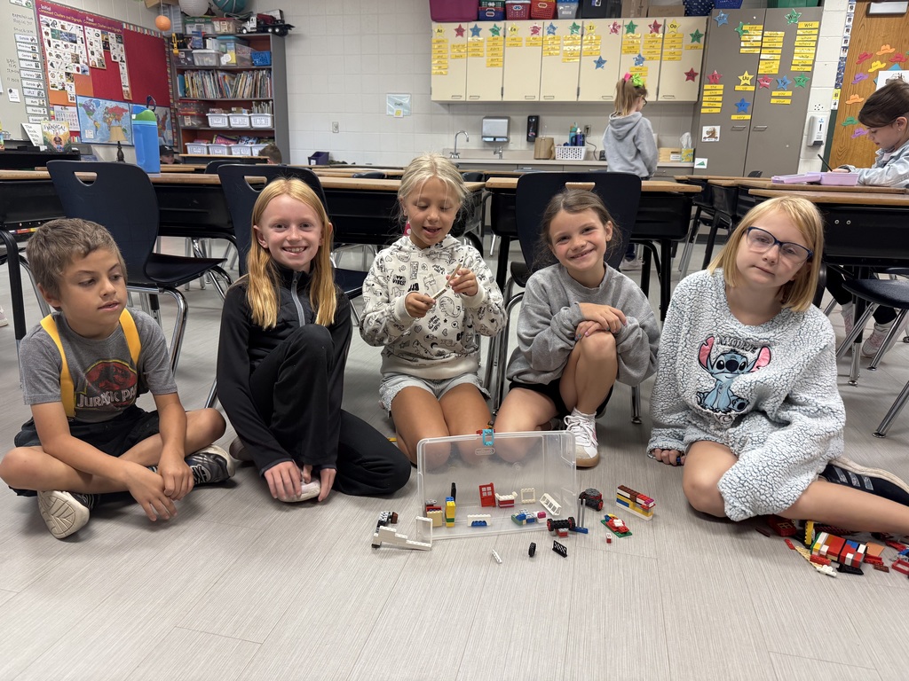 Five students sit on the classroom floor around a bin and scattered LEGO pieces, displaying their build in front.