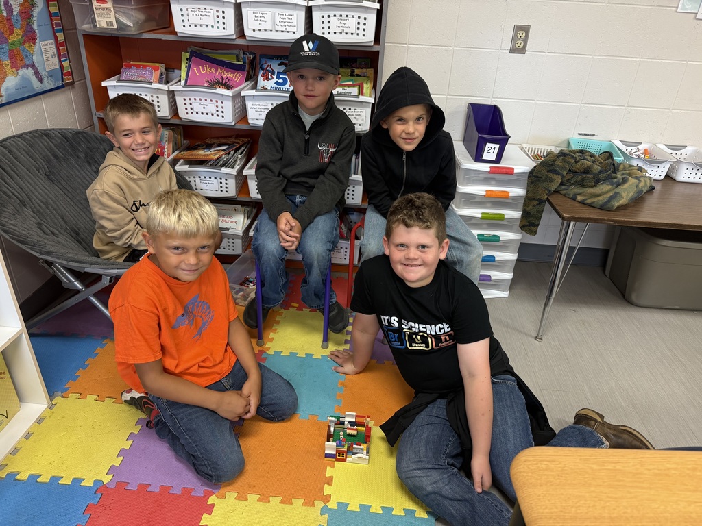 Five students sit on a colorful rug, smiling as they pose with their group LEGO project.