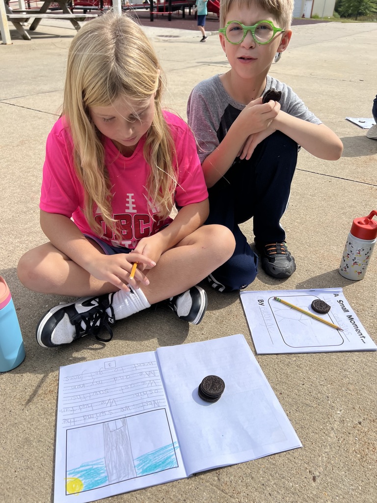 Two students sit on the pavement with writing notebooks open. One child writes while the other eats an Oreo cookie.