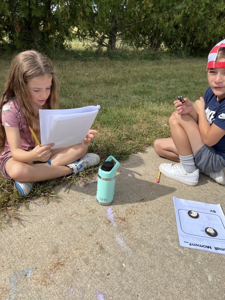 Two students sit outdoors on the pavement with notebooks and worksheets in front of them. One child reads, and the other eats an Oreo cookie.