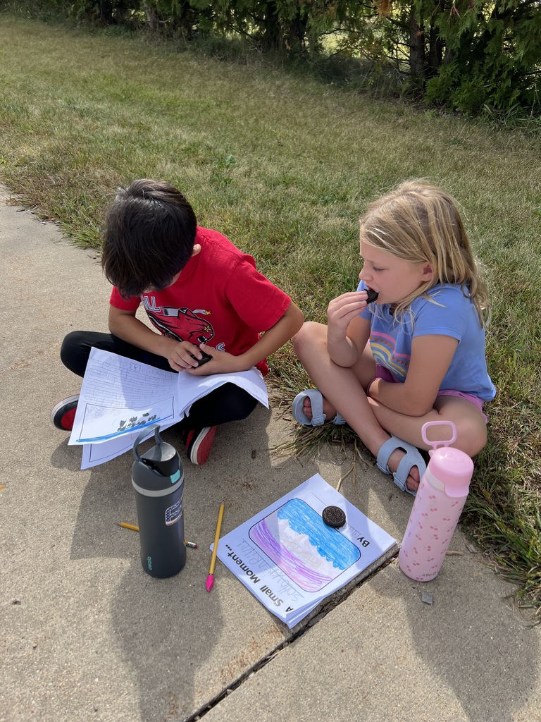 A boy and girl sit on the ground with notebooks and worksheets. The girl eats an Oreo while the boy reads.
