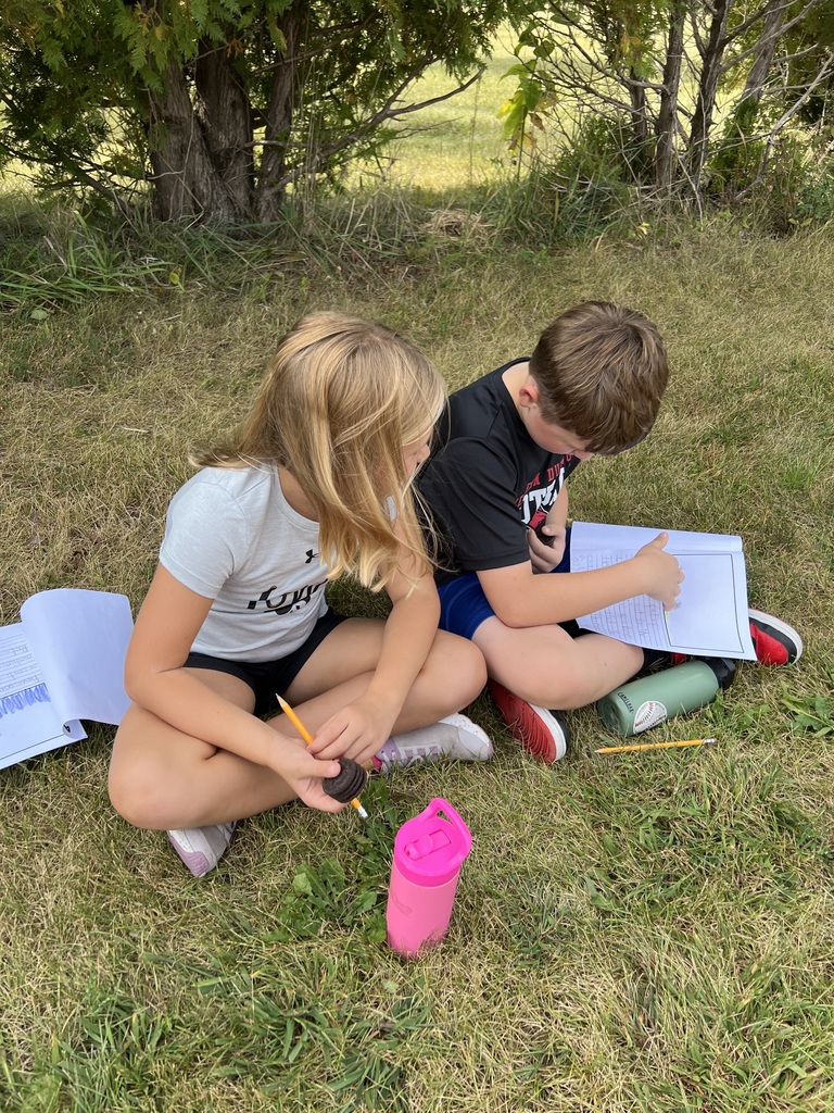 A girl and boy sit on the grass with notebooks open. The girl holds a cookie while the boy writes.