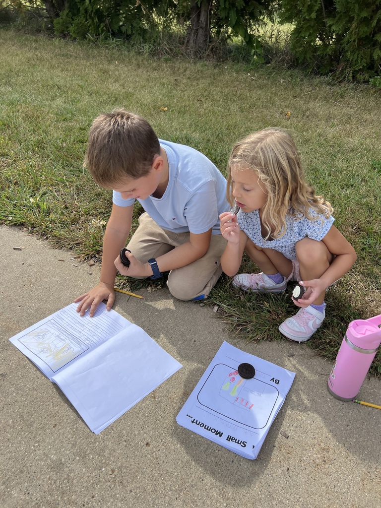 Two students work side by side on the sidewalk with writing journals, each holding an Oreo cookie.