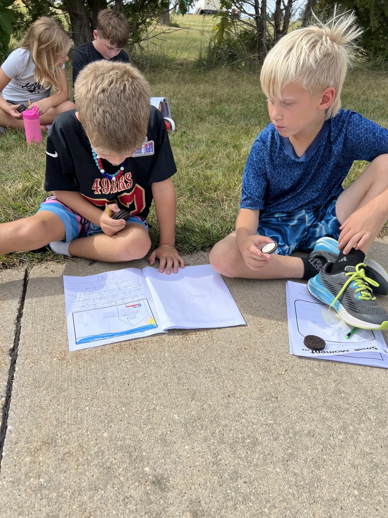 Two boys sit on the grass with their notebooks open, holding Oreo cookies as part of their writing activity.