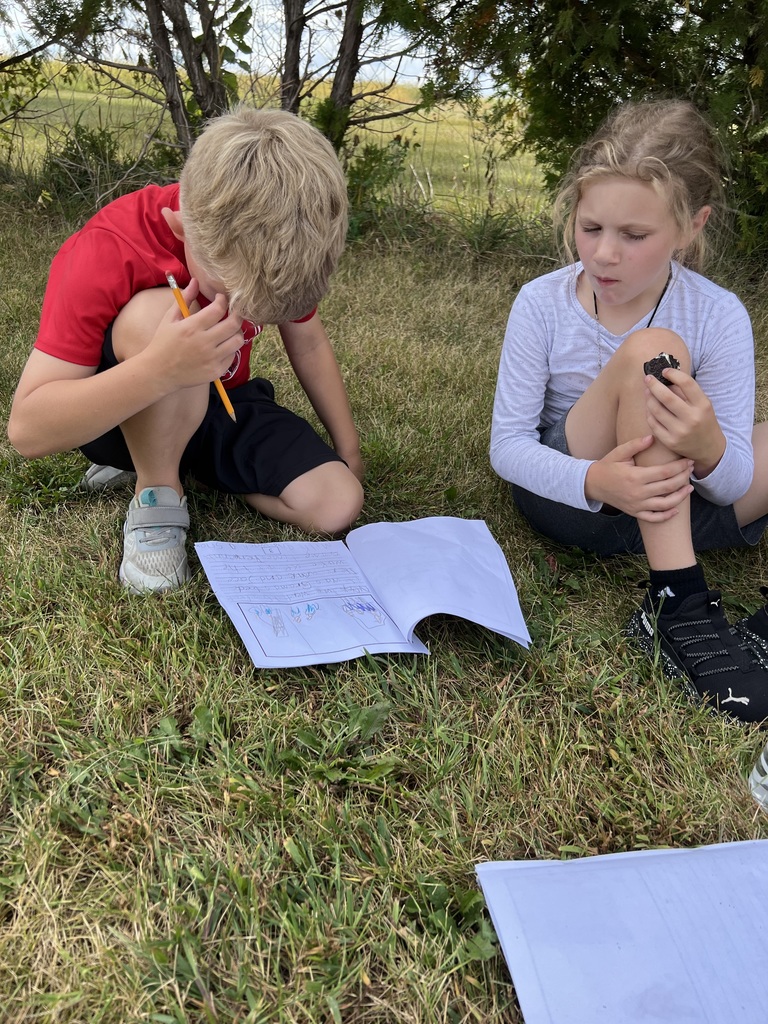 A boy and girl sit in the grass with open notebooks. The girl eats an Oreo while the boy studies his work.