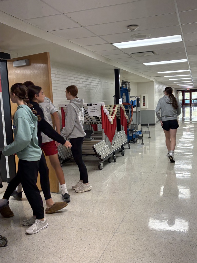 A group of middle school students walks down a school hallway toward an open classroom door. One student is walking ahead while others enter the room together, passing stacked athletic hurdles on carts.