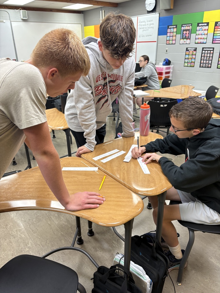 Three students stand around a cluster of desks working on an assignment. One student writes on a paper strip while the others watch and discuss.