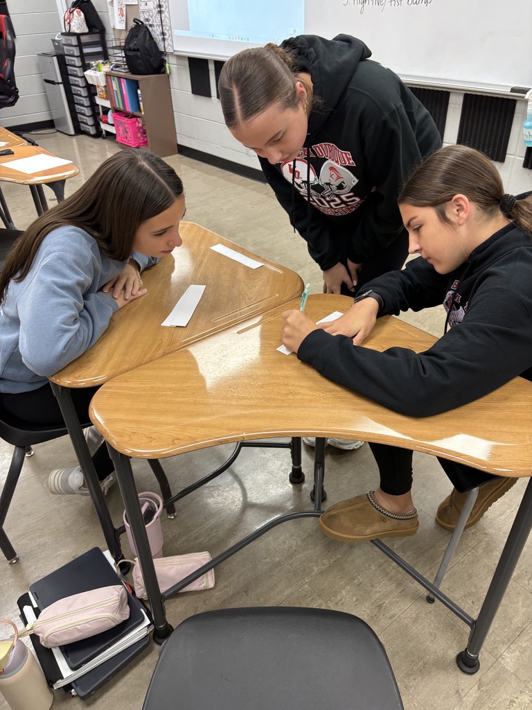 Two students sit at a desk while another stands nearby, leaning in to look at their work. One student writes on a paper strip as the others watch and talk through the activity.