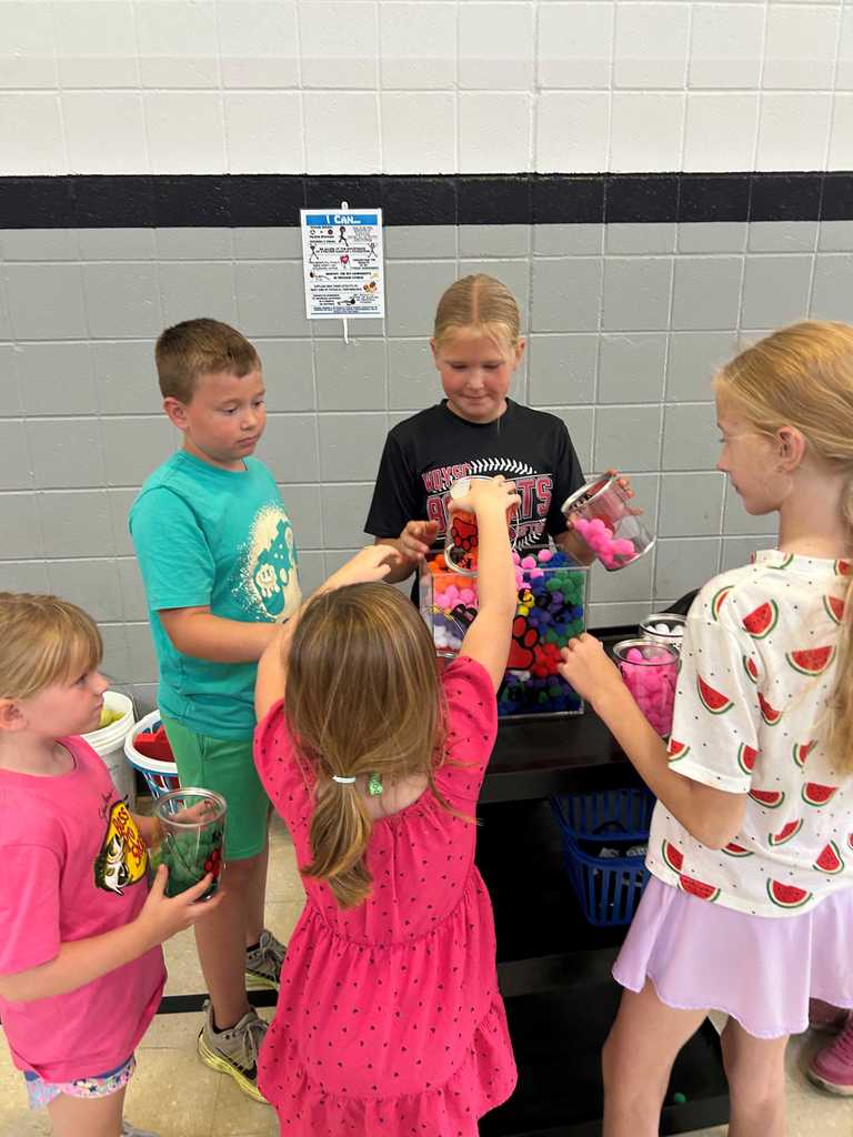 Five children gather around a table, excitedly filling jars with colorful pom-poms.